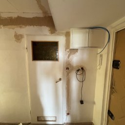 Amper-Wolt - Wnętrze korytarza w trakcie remontu z widoczną białą szafką elektryczną, niebieskim przewodem, domofonem oraz odsłoniętymi puszkami i okablowaniem w ścianie. Tynk na ścianach i suficie jest...