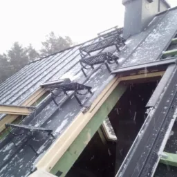 Montaż okna dachowego na pokrytym śniegiem, blaszanym dachu z zamontowanymi stopniami kominiarskimi i widocznym kominem.