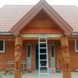 Drewniany dom z rzeźbionymi kolumnami przy wejściu, na tle dachu z brązowej dachówki, drabina oparta o wejście, widoczne okna i drzwi w kolorze jasnoniebieskim.