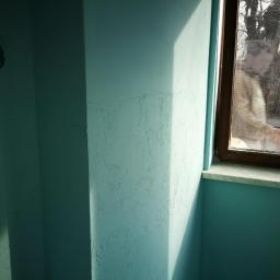 Gładzie Maszynowe Przemysław Kowalski - Narożnik pokoju pomalowany na turkusowo, z widoczną fakturą na ścianie i otworem na instalację elektryczną. W oknie odbija się sylwetka osoby trzymającej zielony przedmiot.