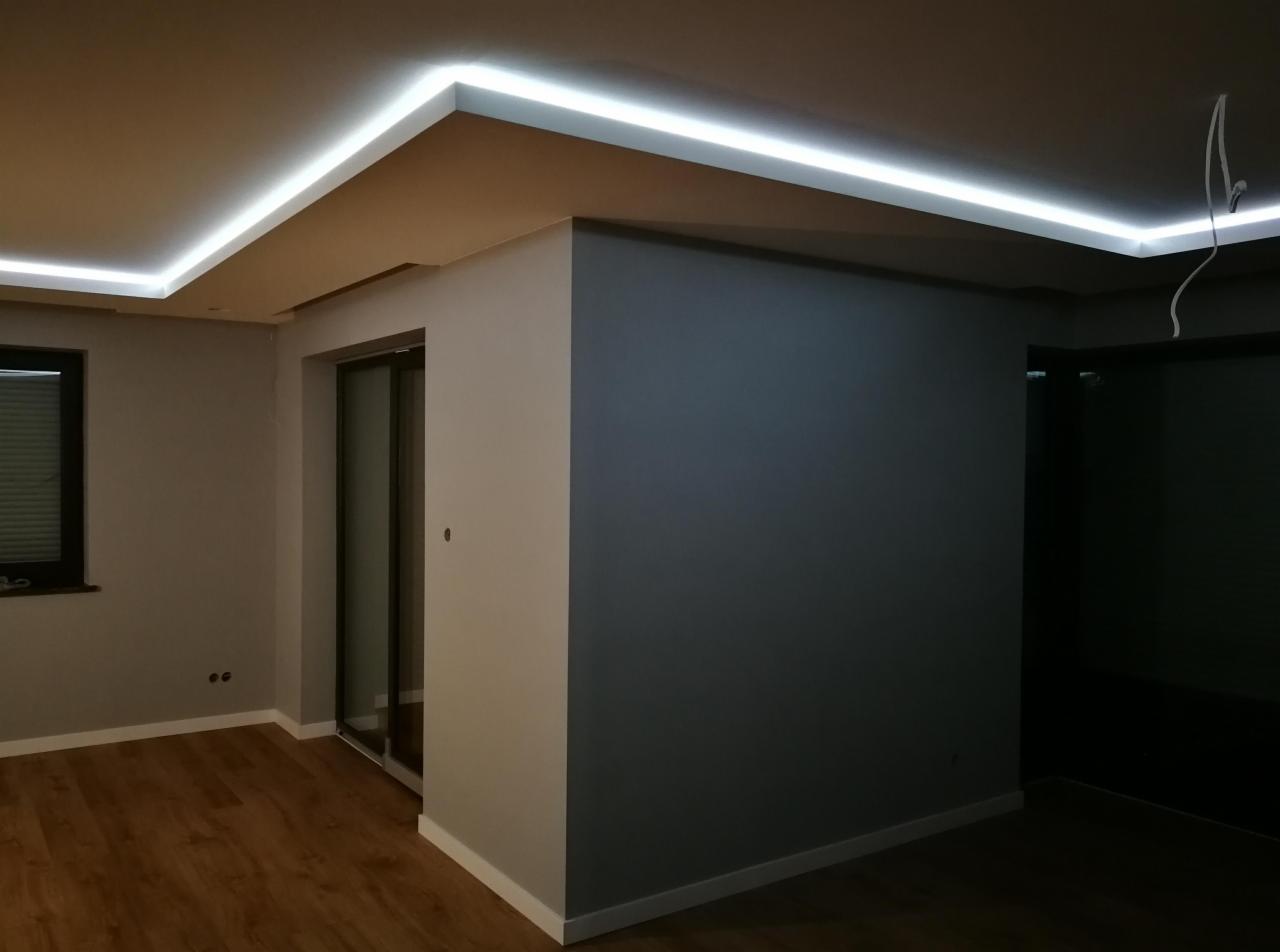 Nowoczesne wnętrze z podwieszanym sufitem i oświetleniem LED, szare ściany i drewniana podłoga, widoczne przewody elektryczne.