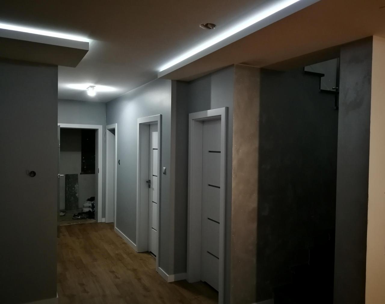 Korytarz w szarościach z drewnianą podłogą i oświetleniem LED w suficie podwieszanym, widoczne trzy białe drzwi z pionowymi frezami, ściany wykończone farbą w odcieniach szarości.