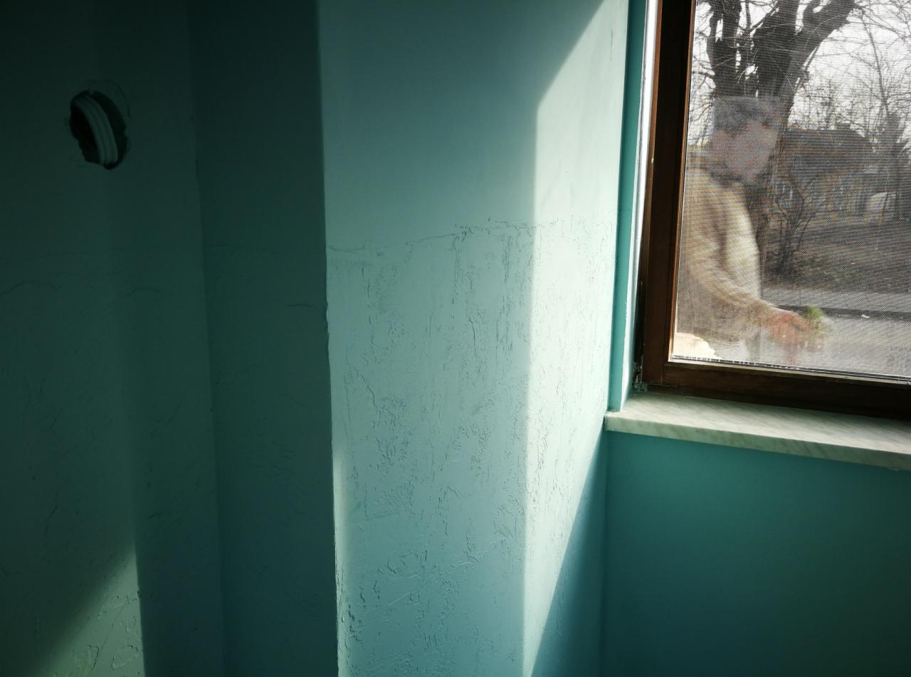 Narożnik pokoju pomalowany na turkusowo, z widoczną fakturą na ścianie i otworem na instalację elektryczną. W oknie odbija się sylwetka osoby trzymającej zielony przedmiot.