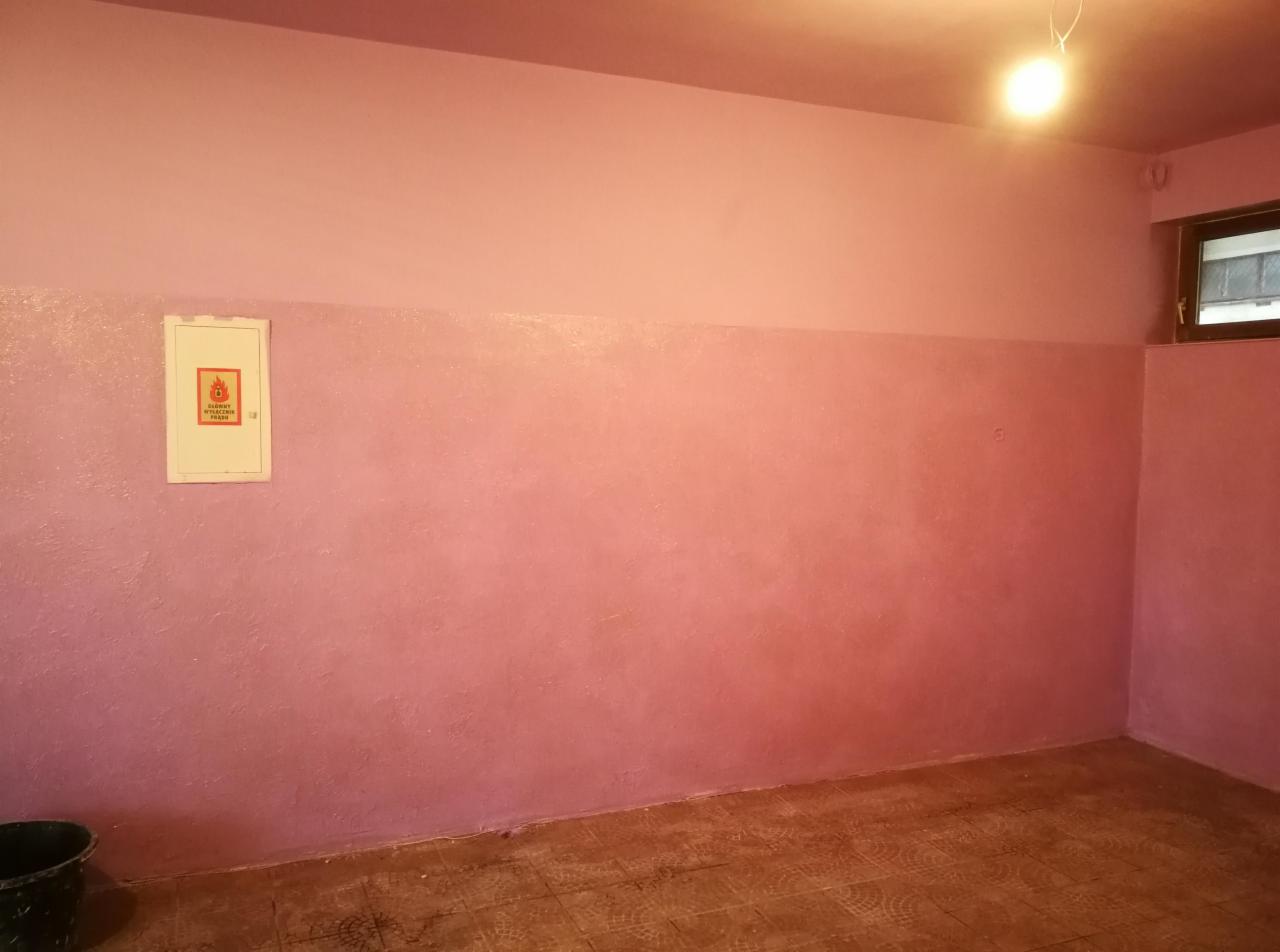 Różowe ściany w pokoju, dolna część ściany z widoczną teksturą, górna część gładka, na ścianie skrzynka elektryczna z piktogramem ognia.