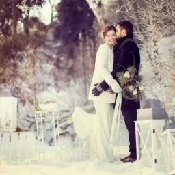 Uśmiechnięta panna młoda w białym płaszczu i pan młody w czarnym płaszczu stojący w zimowej scenerii, dekoracje ślubne z lampionami i drewnianymi elementami w tle.