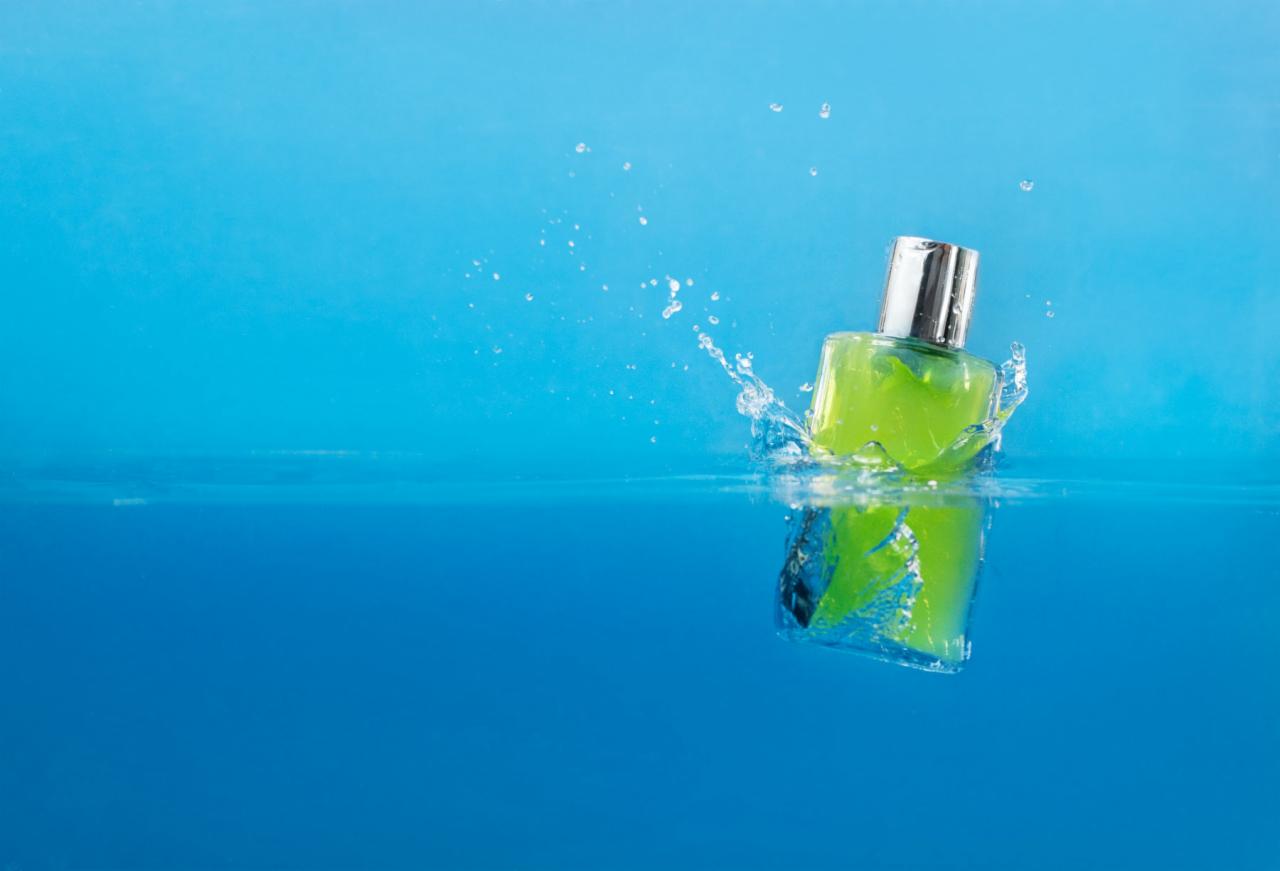 Flakon zielonego perfumu z srebrną nakrętką wpada do wody, tworząc rozprysk na tle błękitnego tła.