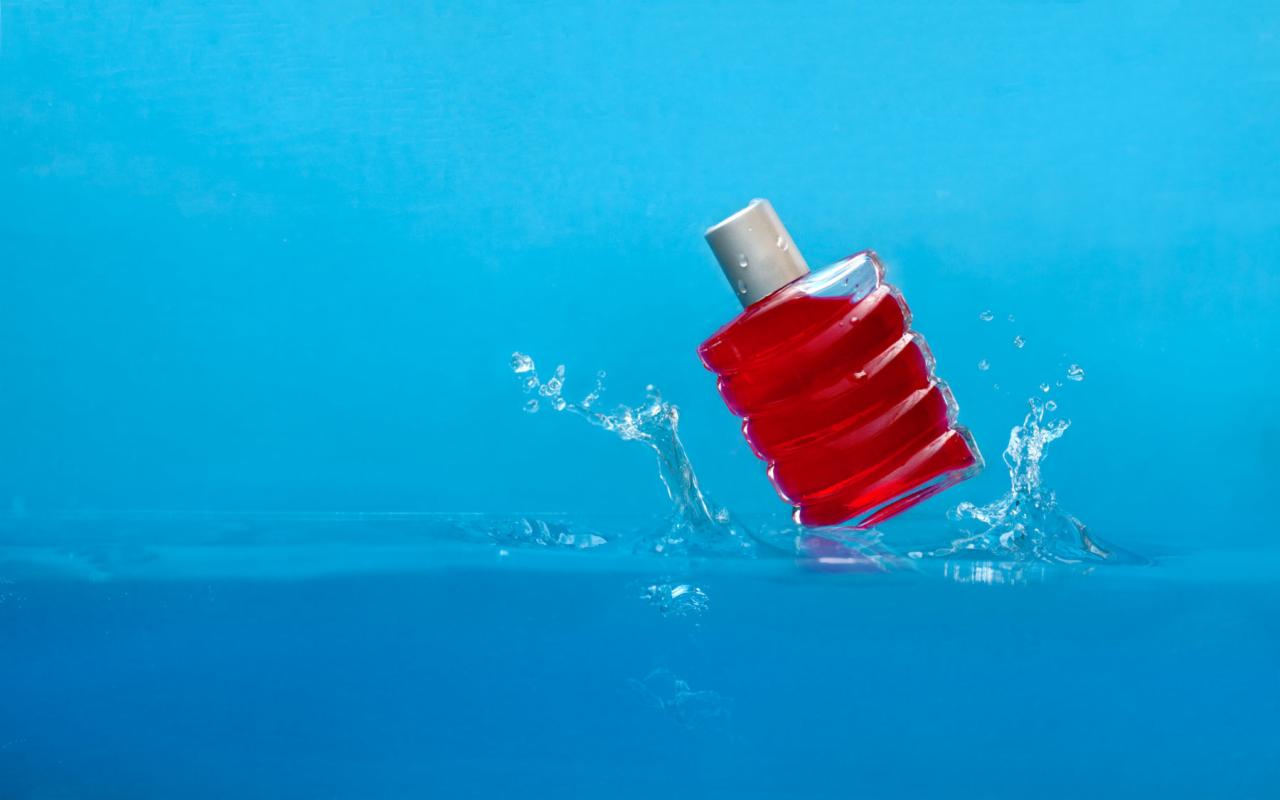 Czerwony flakon perfum z srebrnym korkiem, częściowo zanurzony w wodzie, tworzący dynamiczne rozpryski na tle błękitnego tła.