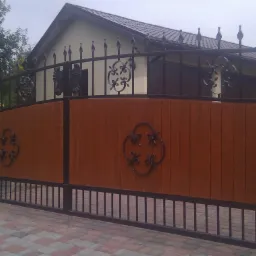 Dwuskrzydłowa brama wjazdowa z drewnianym wypełnieniem i kutymi zdobieniami, osadzona na kamiennych słupkach z kulami, widok na dom z ciemnym dachem.