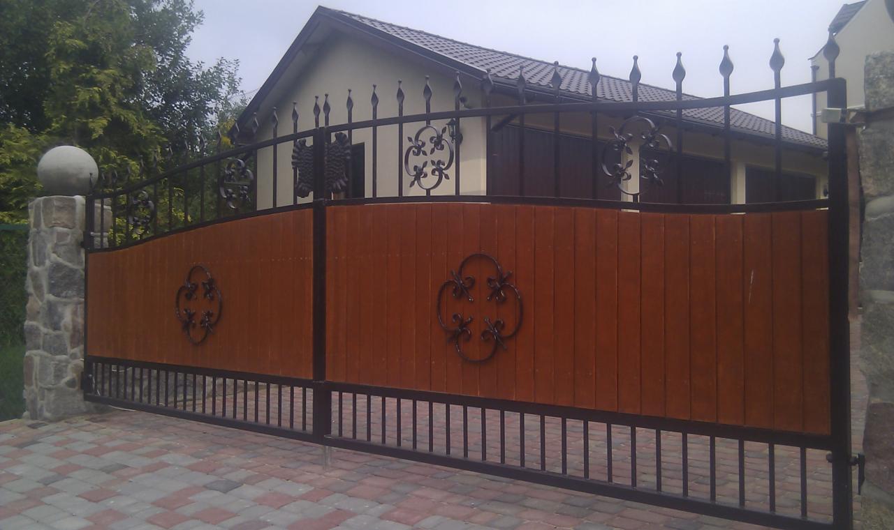 Dwuskrzydłowa brama wjazdowa z drewnianym wypełnieniem i kutymi zdobieniami, osadzona na kamiennych słupkach z kulami, widok na dom z ciemnym dachem.