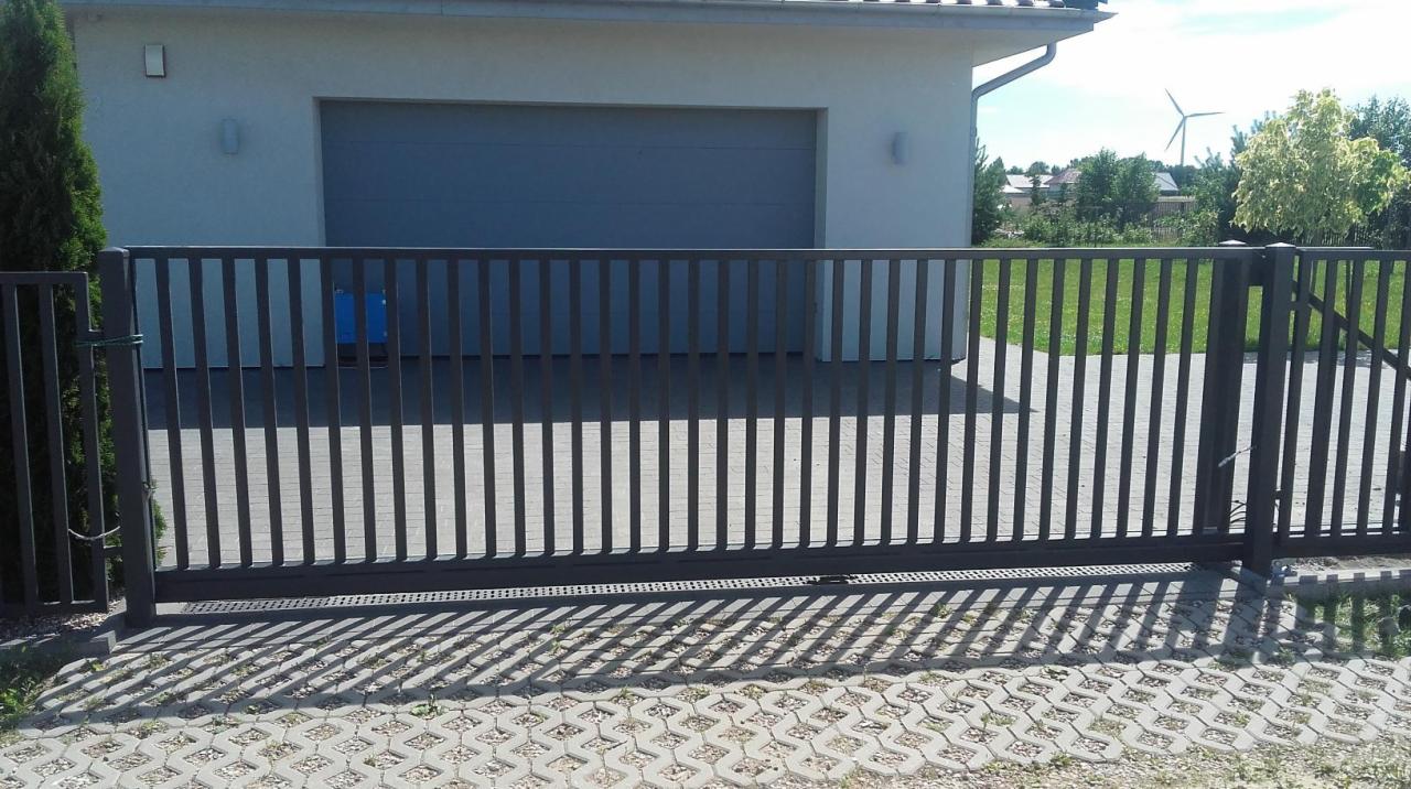Nowoczesna, przesuwna brama wjazdowa z pionowych profili, zautomatyzowana, z widocznym napędem, na tle domu z garażem i turbiną wiatrową w oddali.