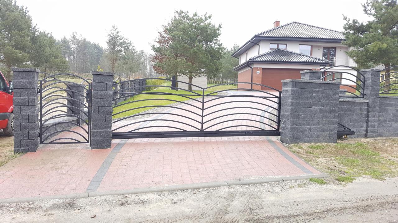 Czarna, przesuwna brama wjazdowa z dekoracyjnymi łukami, osadzona na słupkach z szarej kostki, z widocznym fragmentem ogrodzenia o podobnym wzorze i nowym domem w tle.