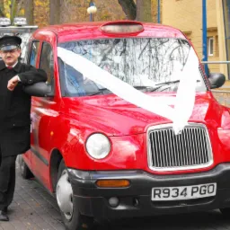 Kierowca w czarnym uniformie opiera się o czerwonego, udekorowanego białą wstęgą taksówkarza, zaparkowanego przed budynkiem.