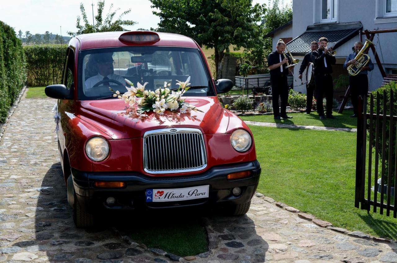 Czerwony samochód typu taxi udekorowany białymi kwiatami na masce, orkiestra dęta w tle, tablica rejestracyjna z napisem 'Młoda Para'.