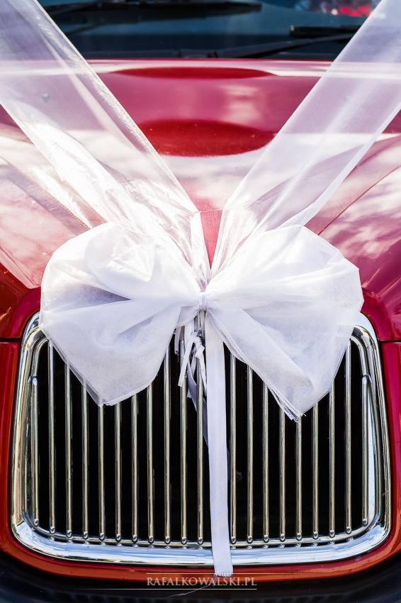 Czerwony samochód udekorowany dużą, białą, tiulową kokardą na masce, widok z przodu na chromowaną atrapę chłodnicy.