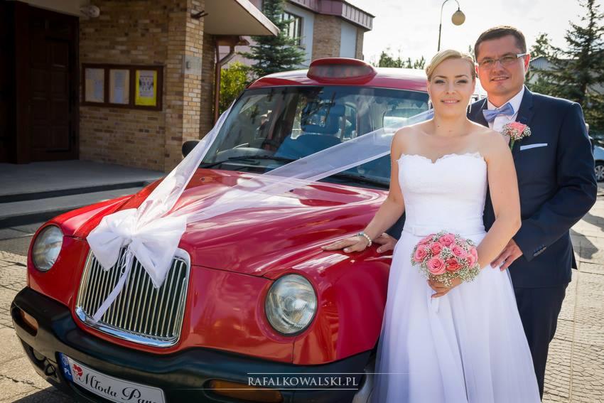 Para młoda pozuje przy czerwonym samochodzie typu londyńska taksówka udekorowanym białą wstążką i kokardą, z tablicą rejestracyjną 'Młoda Para' na tle budynku z cegły.