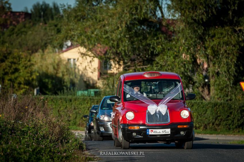 Czerwony, udekorowany kokardami samochód typu taksówka na drodze, z tablicą rejestracyjną 'Młoda Para', w tle zieleń i zabudowania.
