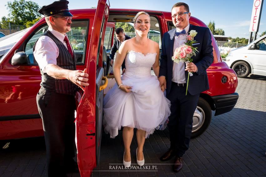 Para młoda wysiadająca z czerwonej taksówki z szoferem w kapeluszu i kamizelce, trzymającym drzwi.