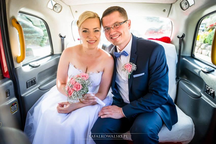 Uśmiechnięta para młoda w taksówce: panna młoda w białej sukni trzyma bukiet różowych róż, pan młody w granatowym garniturze z błękitną muchą i różową butonierką.
