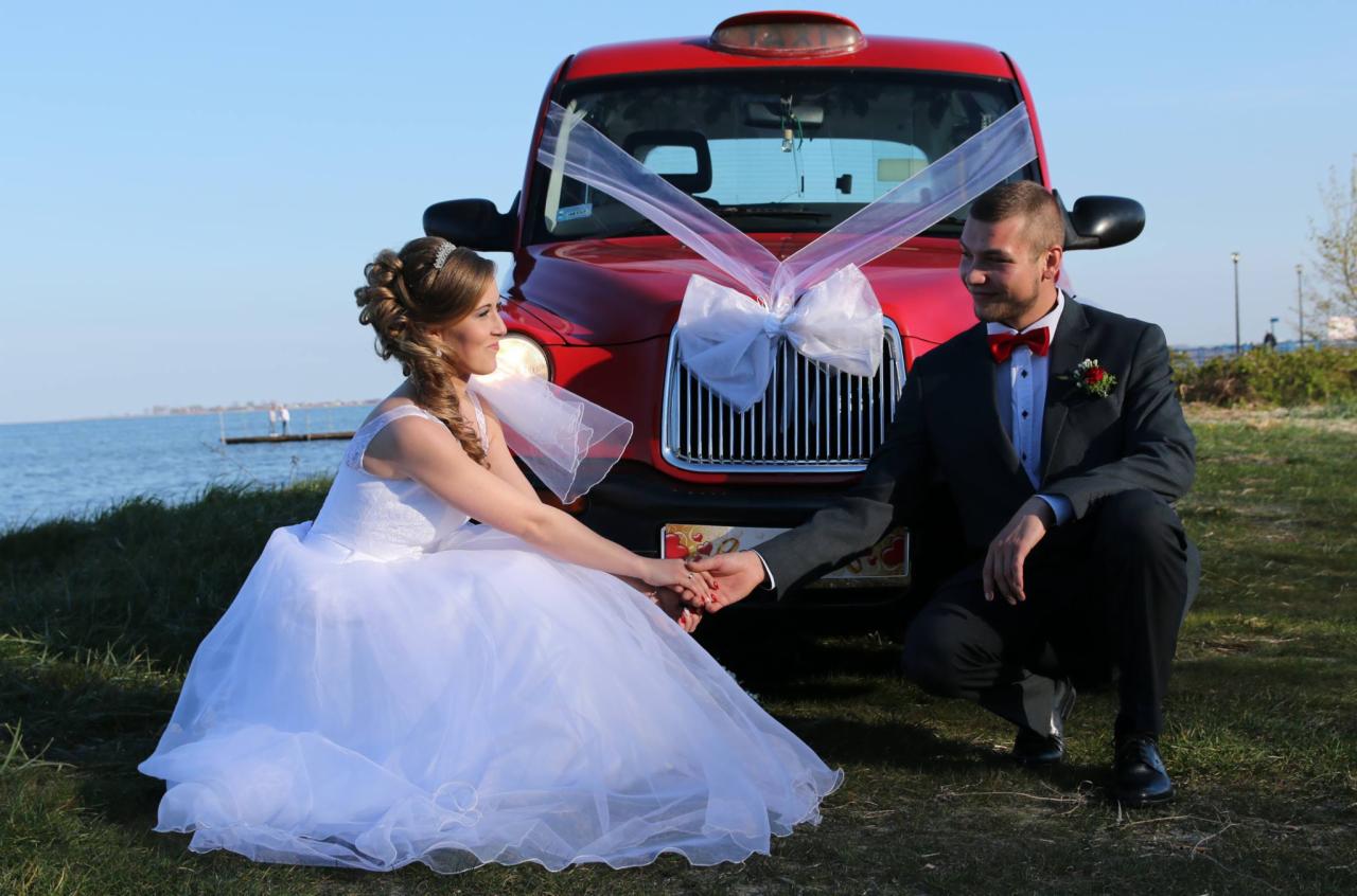 Para młoda trzymająca się za ręce przed czerwonym samochodem udekorowanym białymi kokardami, w tle jezioro i niebieskie niebo.