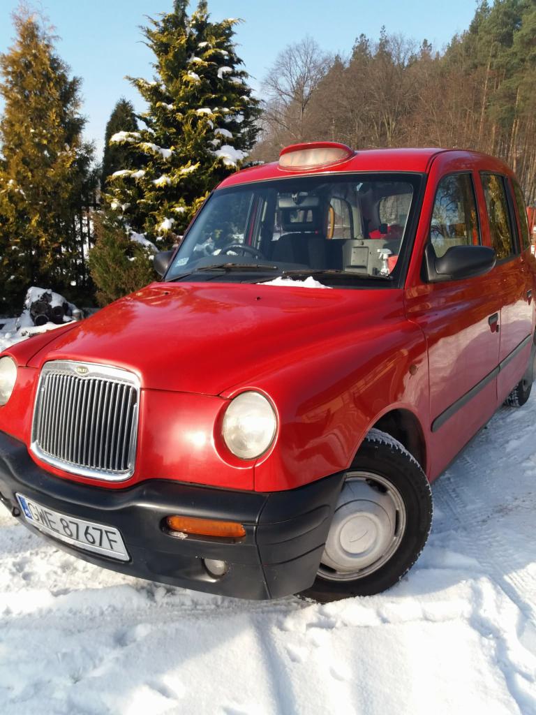Czerwona taksówka w stylu londyńskim, zaparkowana na śniegu w zimowej scenerii, z widocznymi drzewami w tle.
