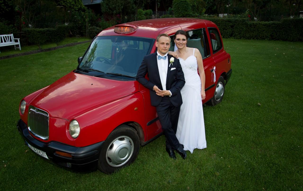Para młoda w strojach ślubnych pozuje przy czerwonym samochodzie typu taksówka na trawniku.
