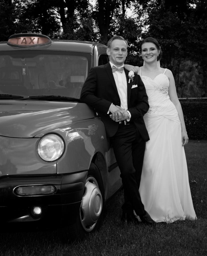 Para młoda w strojach ślubnych pozuje przy samochodzie taxi w stylu retro, czarno-białe zdjęcie.