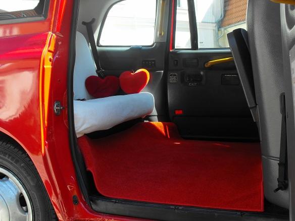 Czerwony samochód z otwartymi drzwiami, wnętrze z czerwonym dywanem i białym siedzeniem ozdobionym dwoma czerwonymi poduszkami w kształcie serc.