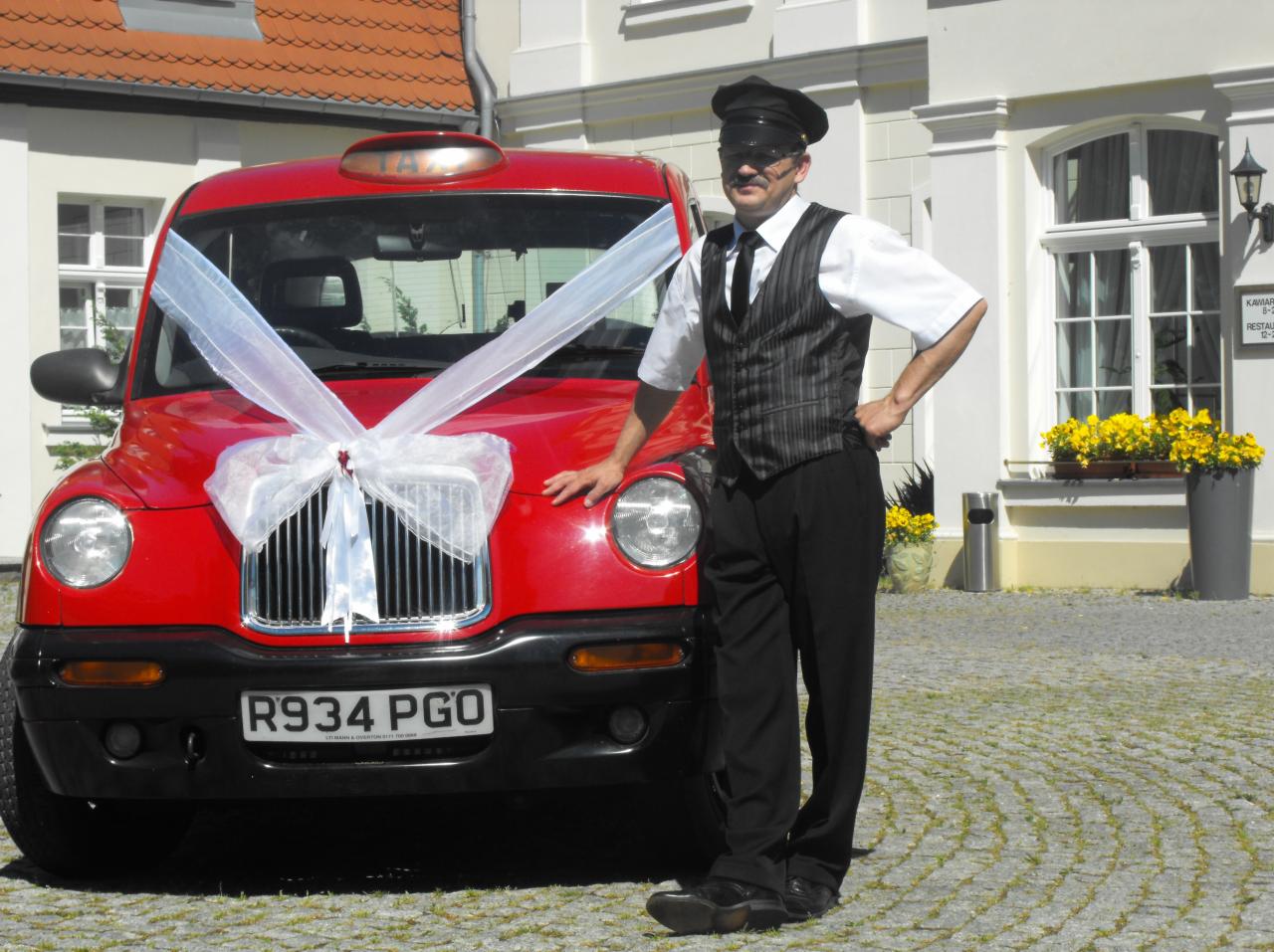 Kierowca w uniformie stoi obok czerwonej taksówki London Taxi udekorowanej białą wstążką, na tle budynku z oknami i kwiatami.