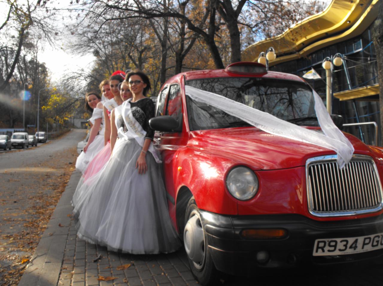 Grupa młodych kobiet w sukniach balowych i szarfach stoi obok czerwonego samochodu typu taksówka, udekorowanego białą wstęgą, zaparkowanego na brukowanej ulicy.