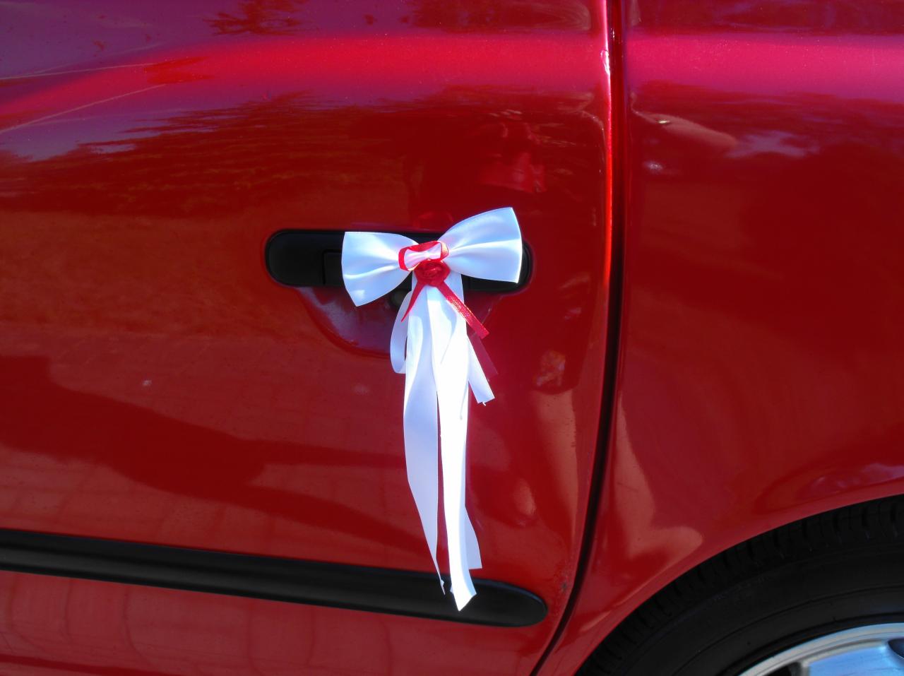 Czerwony samochód udekorowany białą kokardą z czerwonym akcentem na klamce, prawdopodobnie przygotowany do ślubu.