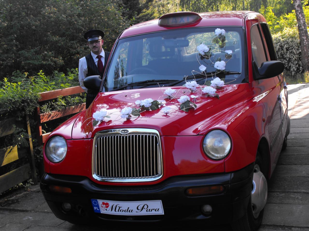 Czerwony, udekorowany białymi kwiatami samochód typu taksówka z kierowcą w czapce i kamizelce, przygotowany do ślubu, z tablicą rejestracyjną 'Młoda Para'.