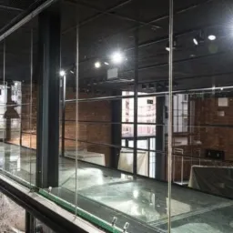 Szklana podłoga i balustrada w loftowym wnętrzu z ceglaną ścianą, widok z góry na konstrukcję z metalowymi ramami i przezroczystym szkłem.