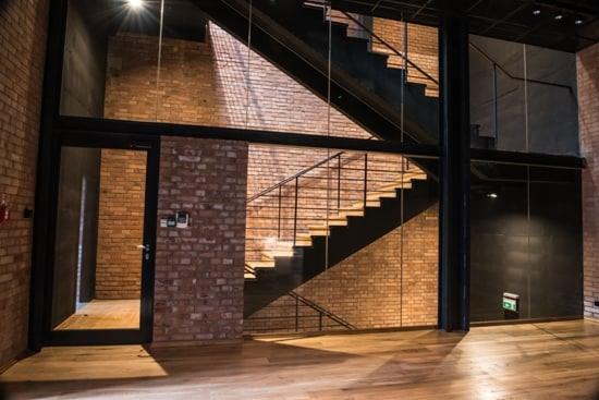 Wnętrze w stylu loft z ceglaną ścianą, drewnianą podłogą i czarną, metalową konstrukcją schodów z drewnianymi stopniami, widoczne przeszklone drzwi z czarną ramą.