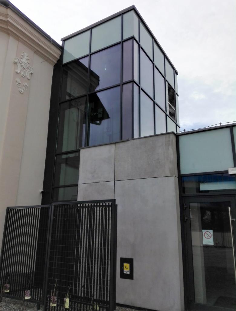 Szklana nadbudowa budynku z ciemnymi ramami okiennymi, widoczna boczna elewacja z dekoracyjnym elementem, czarne metalowe ogrodzenie z przodu, szare tynki.