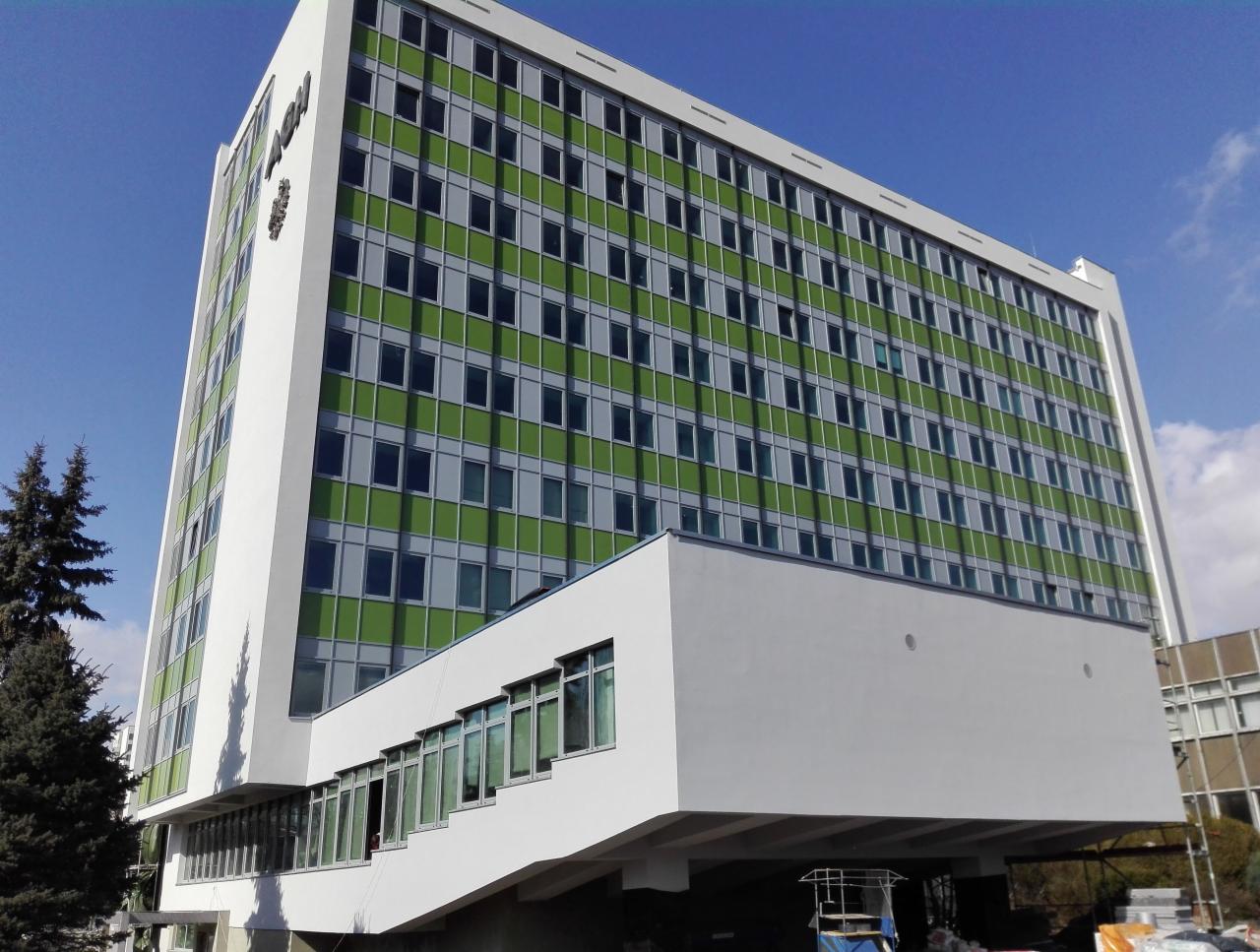 Wysoki budynek biurowy z zielonymi i szarymi oknami, sfotografowany pod kątem z dołu w słoneczny dzień.