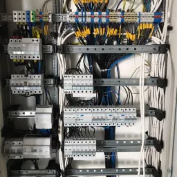 Rozdzielnica elektryczna z licznymi przewodami, zabezpieczeniami i listwami zaciskowymi, starannie uporządkowana w szafie, widoczne oznaczenia kabli.