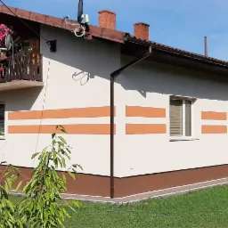 Dwukondygnacyjny dom z jasną elewacją, ozdobiony poziomymi pasami w kolorze terakoty, z balkonem udekorowanym kwiatami, na tle zielonego ogrodu i błękitnego nieba.