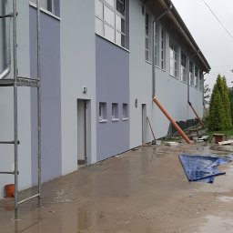 Artur Ciekot - Dwukondygnacyjny budynek po niedawnym malowaniu elewacji, z widocznym rusztowaniem po lewej stronie i mokrym terenem przed budynkiem, sugerującym świeżo zakończone prace w deszczową pogodę.