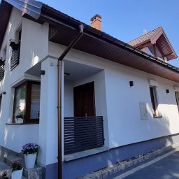 Artur Ciekot - Biały dom jednorodzinny z ciemnobrązowym dachem i rynnami, widok z zewnątrz, z kwiatami w donicach na oknach i przy wejściu, na tle błękitnego nieba i zieleni drzew.