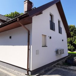 Artur Ciekot - Biały dom jednorodzinny z brązowymi elementami wykończeniowymi, widok z boku na elewację z dwoma klimatyzatorami, kostka brukowa na podjeździe i zielone drzewa w tle.