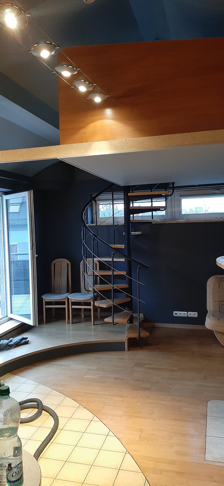 Wnętrze mieszkania w Częstochowie z drewnianą podłogą, spiralnymi schodami z czarną balustradą i drewnianymi stopniami, dwiema krzesłami oraz oknem z widokiem na zewnątrz.