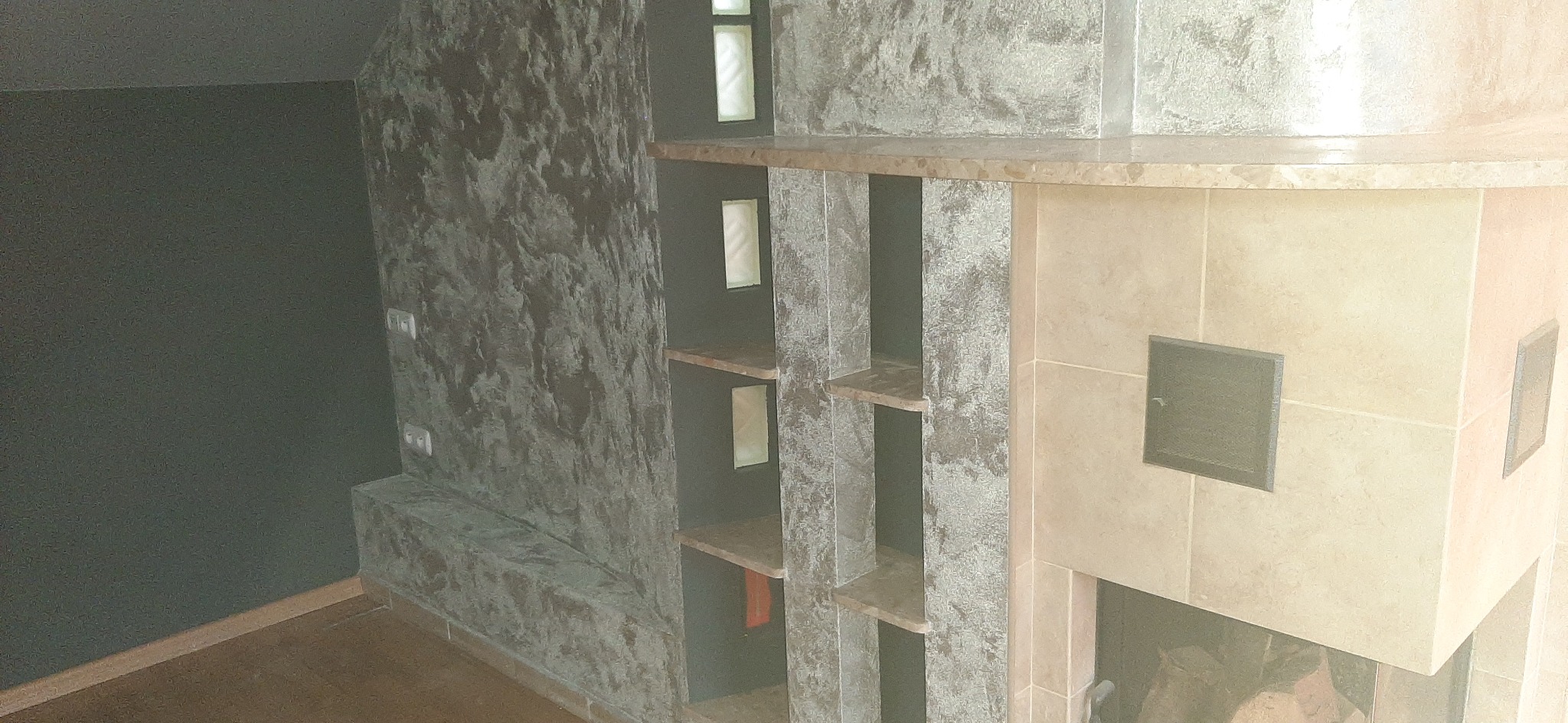 Wykończenie wnętrza z dekoracyjnym tynkiem imitującym kamień na ścianie z półkami i obudową kominka z jasnych płytek ceramicznych.