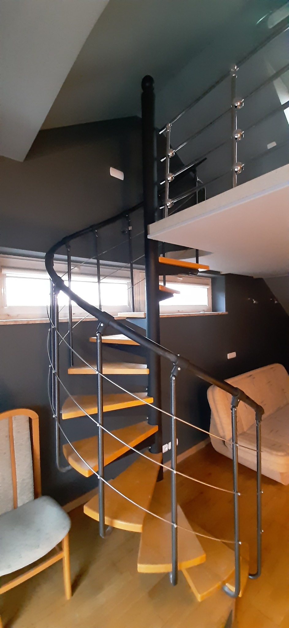 Wewnętrzne spiralne schody z drewnianymi stopniami i czarną metalową konstrukcją, widoczne na tle szarej ściany i okien.