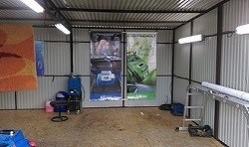 Wnętrze myjni samochodowej z widocznym sprzętem do czyszczenia tapicerki, krzesłem i dwoma banerami reklamowymi na ścianie z blachy falistej, oświetlone lampami jarzeniowymi.