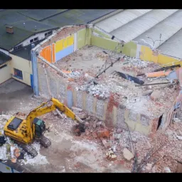 Żółta koparka na tle częściowo zburzonego budynku z kolorowymi ścianami wewnętrznymi. Widok z góry ukazuje gruz i otoczenie budowy.