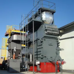 Duży, przemysłowy piec grzewczy z metalowymi schodami i zbiornikiem izolowanym folią aluminiową, widoczny na zewnątrz budynku w słoneczny dzień.