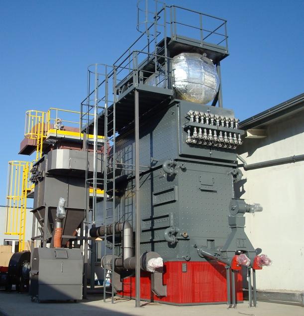 Duży, przemysłowy piec grzewczy z metalowymi schodami i zbiornikiem izolowanym folią aluminiową, widoczny na zewnątrz budynku w słoneczny dzień.