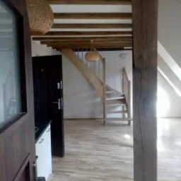 Wnętrze mieszkania po remoncie: widok na drewniane schody, belkowany sufit i lampę z wikliny, z perspektywy otwartych drzwi.