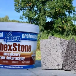 Puszka z mozaikowym tynkiem dekoracyjnym KabexStone na tle próbek tynku i zieleni, prezentacja produktu na budowie lub w składzie budowlanym.