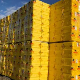 Stosy żółtych płyt izolacyjnych XPS, owiniętych w przezroczystą folię z nadrukowanym logo producenta, ułożone na zewnątrz pod błękitnym niebem.
